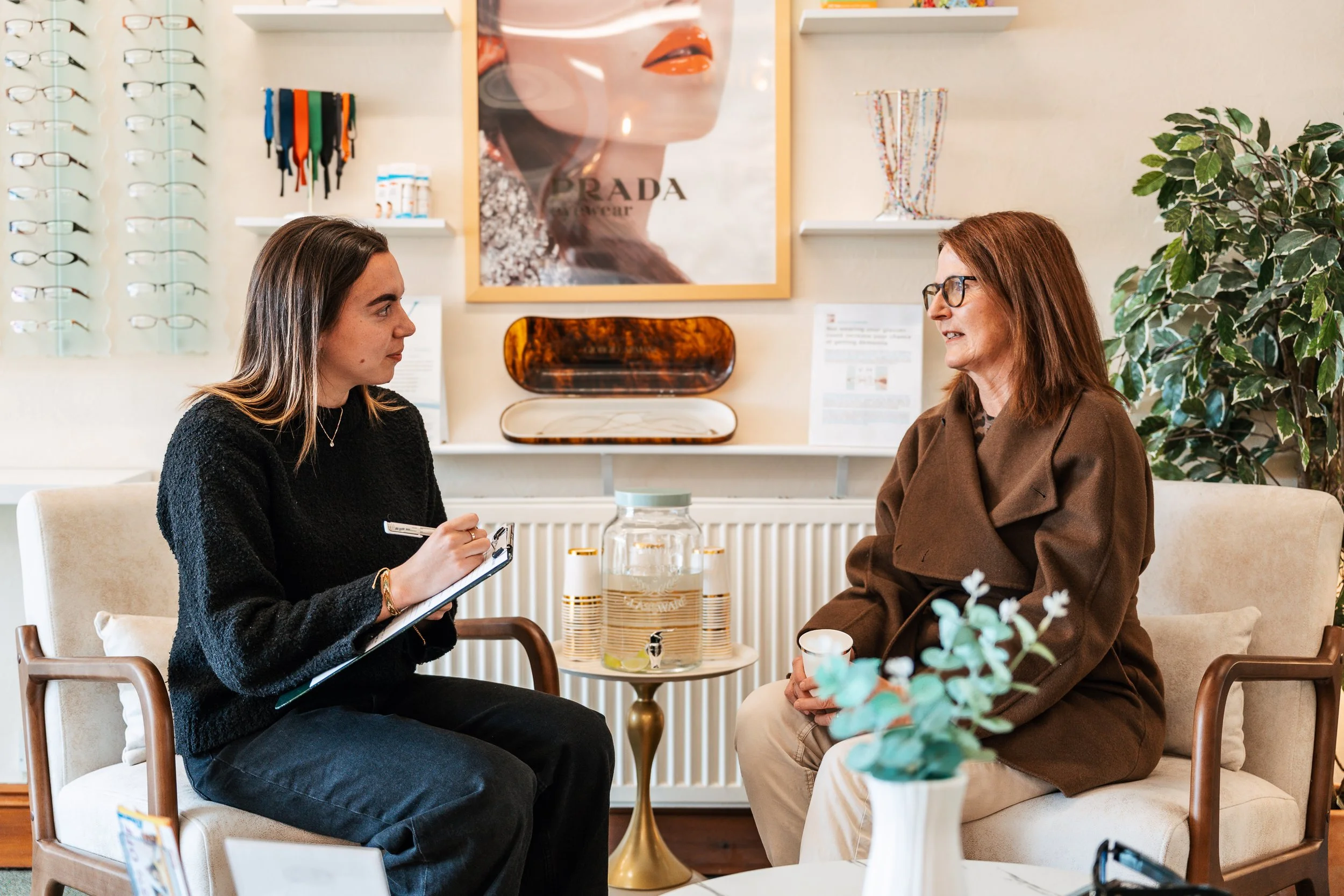 Two women engaged in conversation in a stylish interior, with eyeglasses on display on the wall behind them and a large Prada poster in the background.