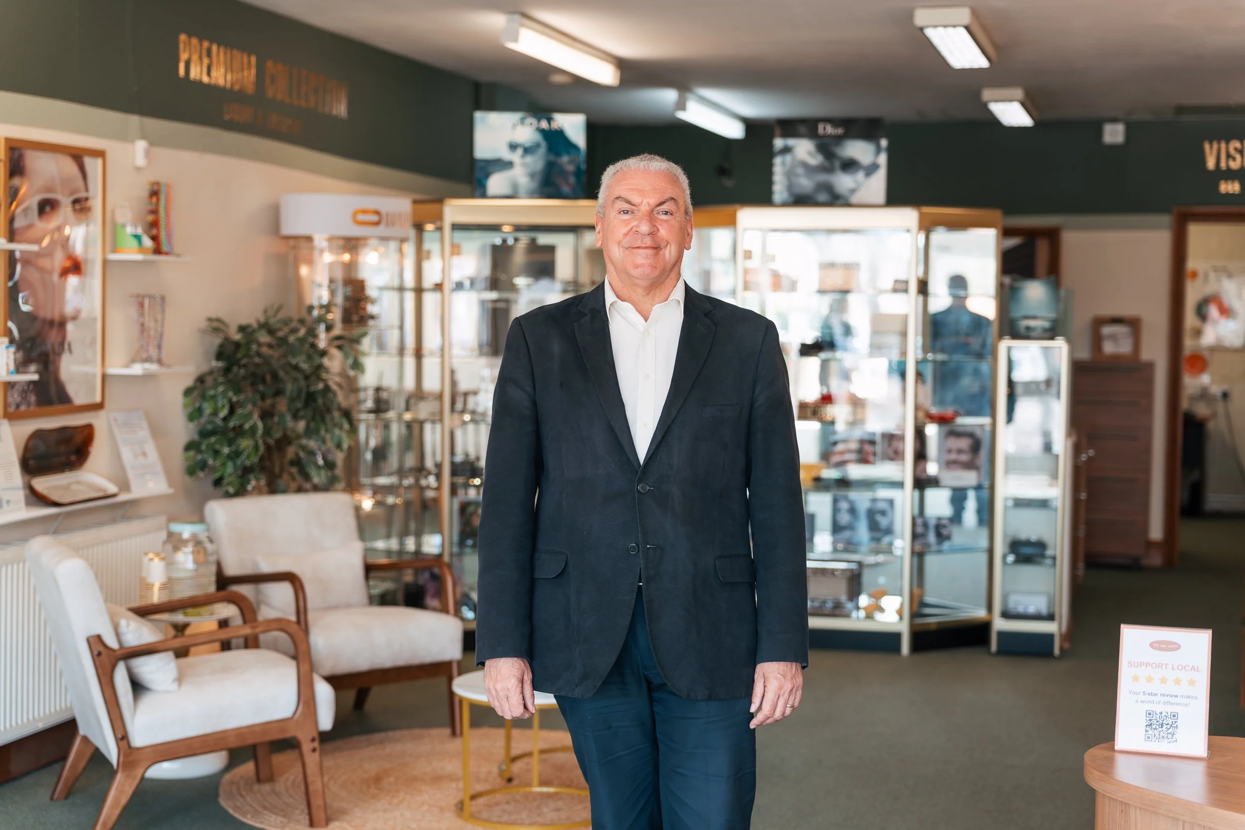 An older man with gray hair, wearing a black blazer and white shirt, standing inside a furniture store with chairs, a plant, and display cases in the background.