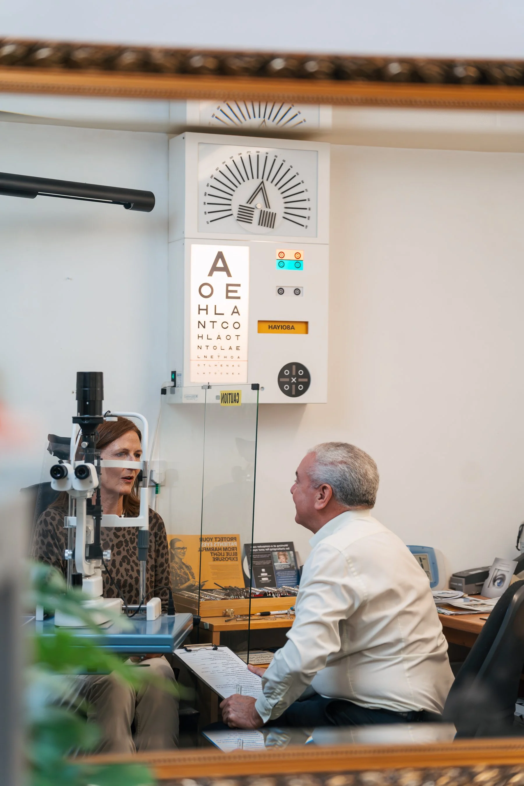 A scene in an optometrist's office showing an eye examination with a woman wearing a leopard print top and a man in a white shirt seated across from each other, with an eye chart, medical equipment, and staff in the background.