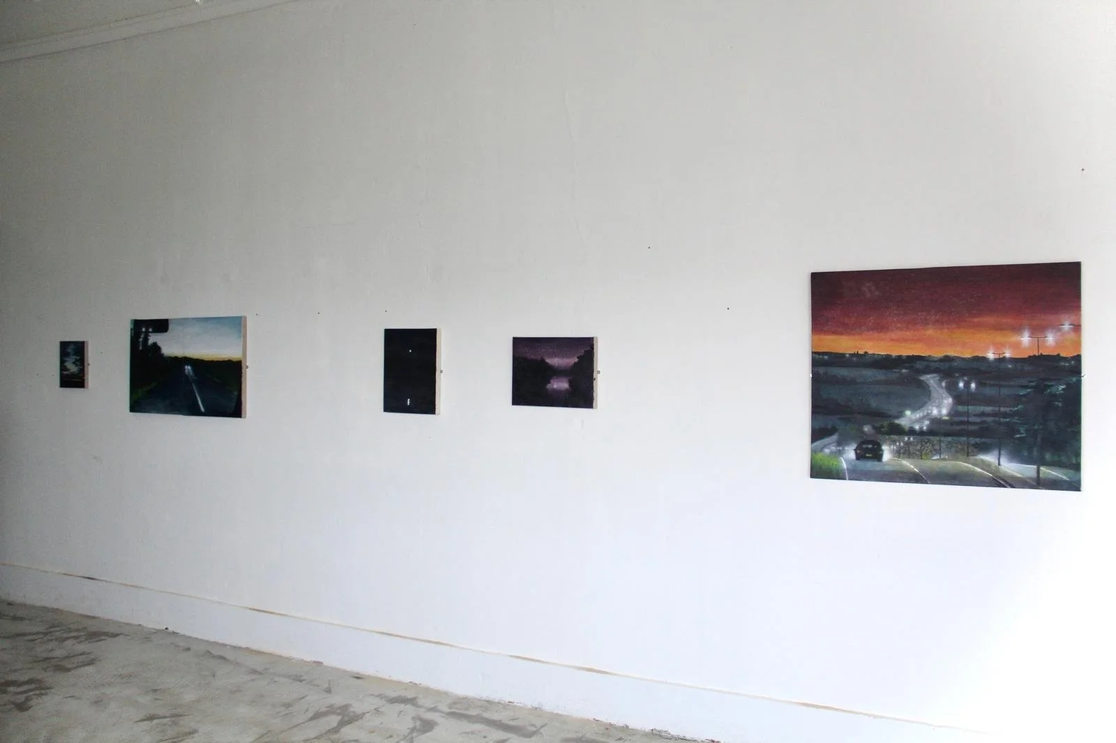 Gallery wall with five landscape photographs of roads and sunsets displayed on a white wall in an art exhibit space.
