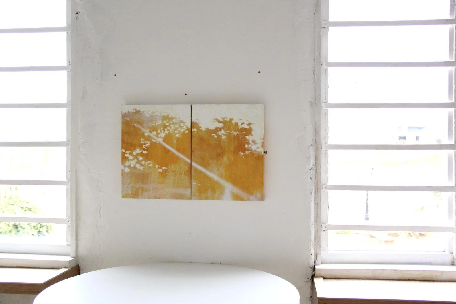 Interior of a room with a white wall, two large windows with horizontal blinds, a round white table in the foreground, and an abstract painting with warm tones hanging on the wall.