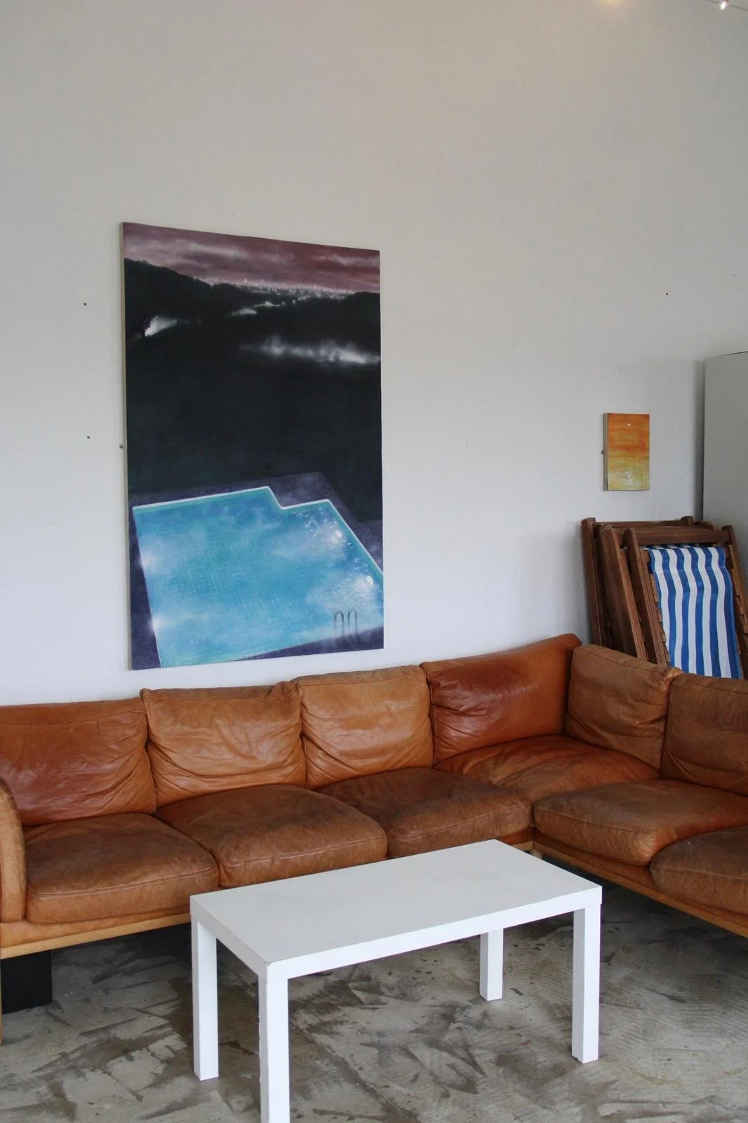 Living room with a tan leather sectional sofa, a white coffee table, wall art including a large painting of a pool and mountains, and a smaller painting of a sunset, with stacked chairs in the background.