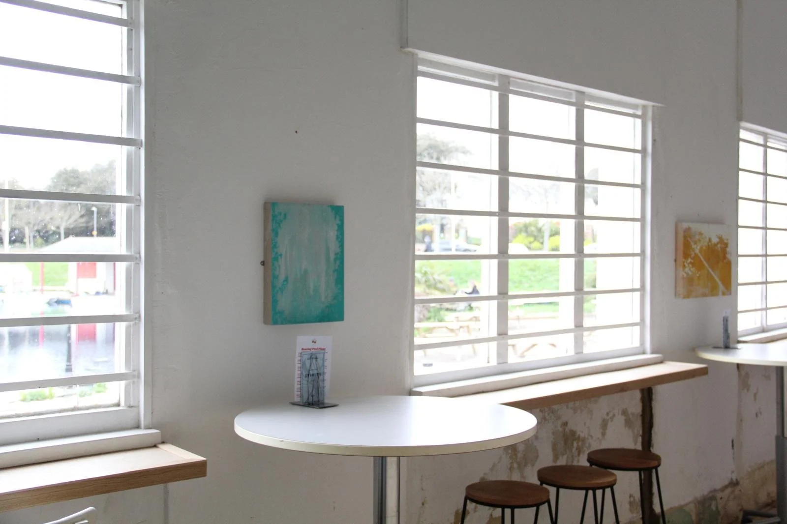 Interior of a cafe or art gallery with white walls, large windows with horizontal white blinds, two small paintings on the walls, and a white round table with three wooden stools underneath. Outside the windows, trees and a sidewalk are visible.