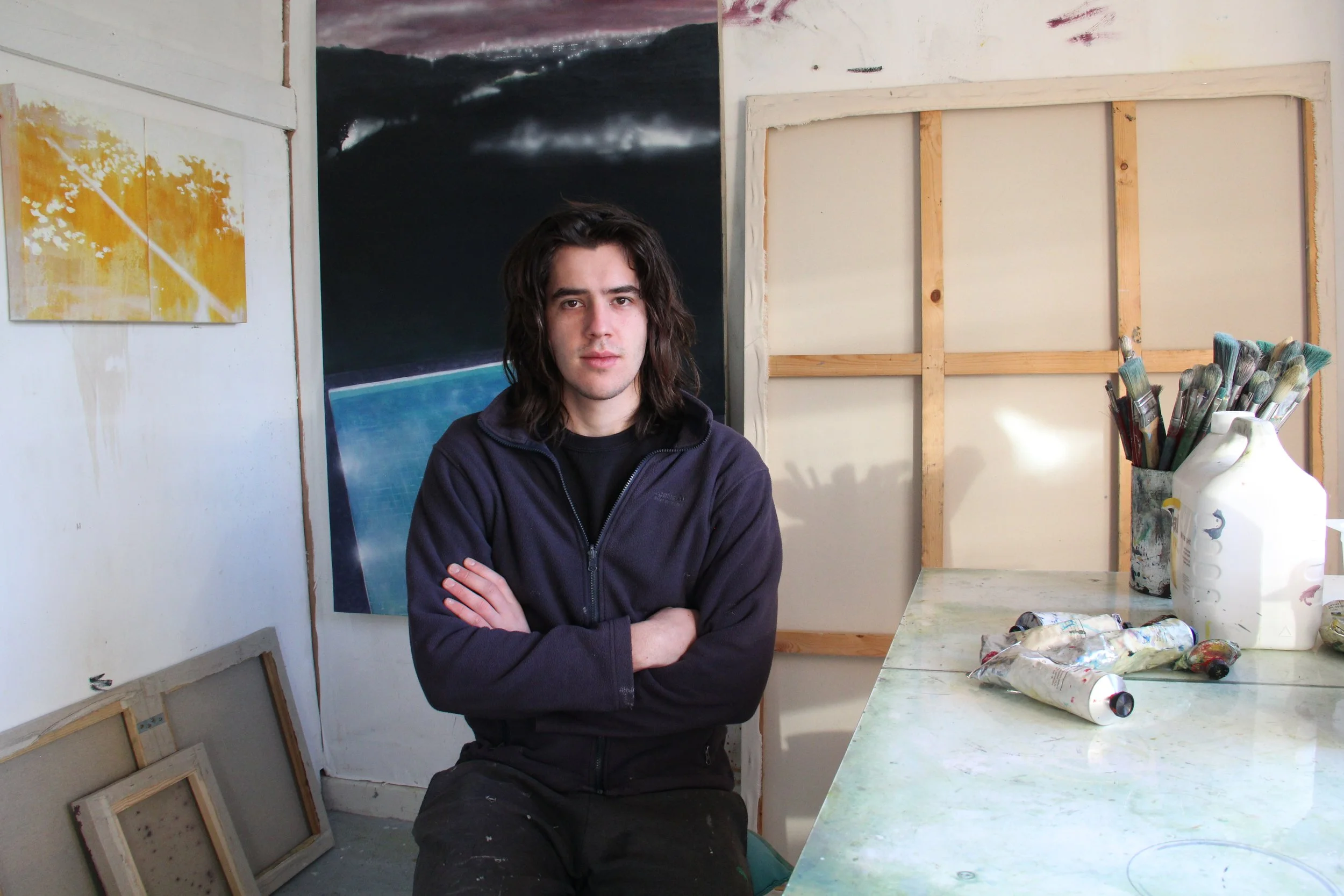 Peter John in an artist's studio, surrounded by paintings and art supplies, including brushes and tubes of paint on a table, surrounded by his work.