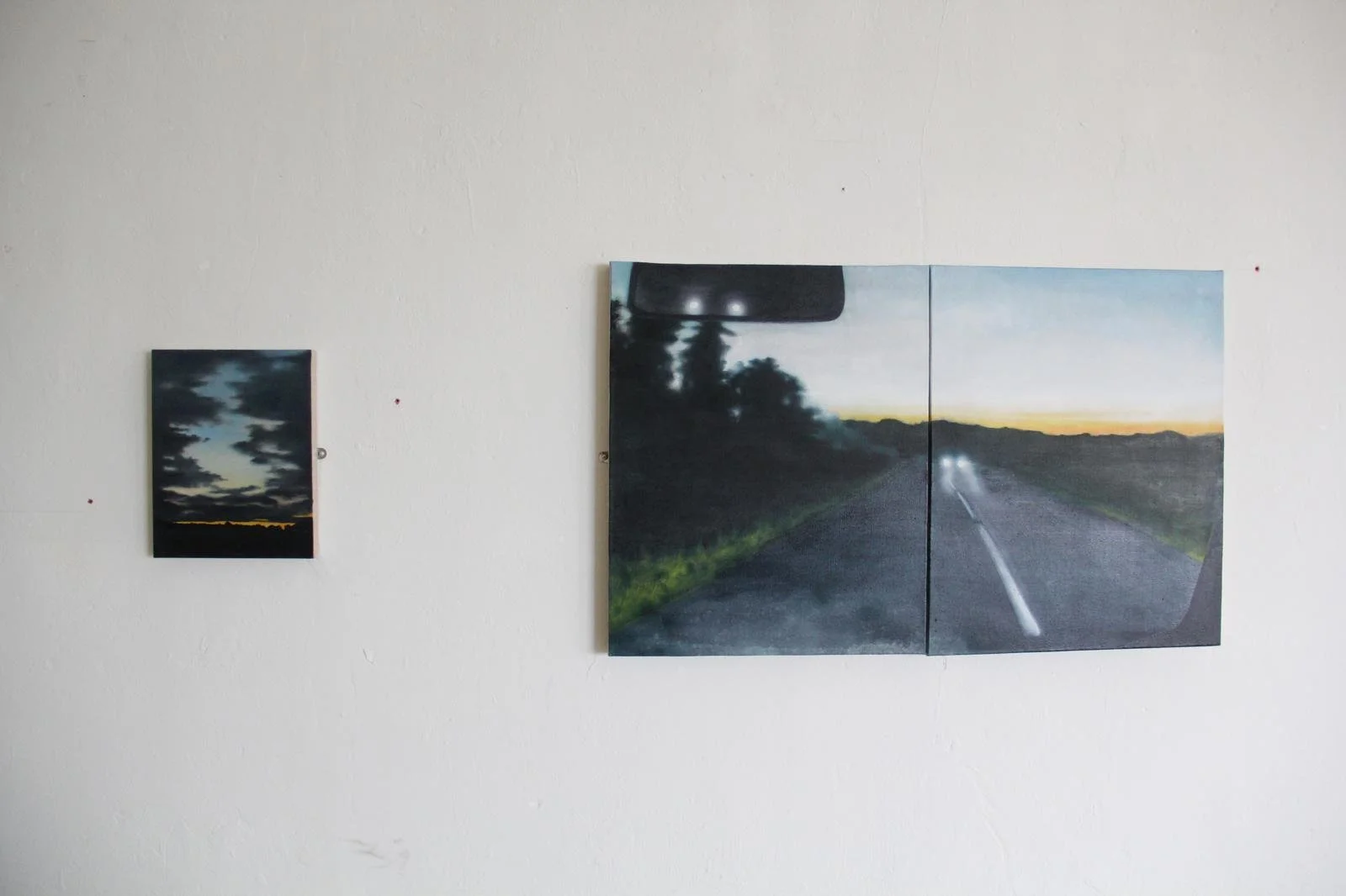 Two landscape paintings on a white wall, one smaller with a cloudy sky at sunset, the other larger depicting a road at dusk with headlights in the distance.
