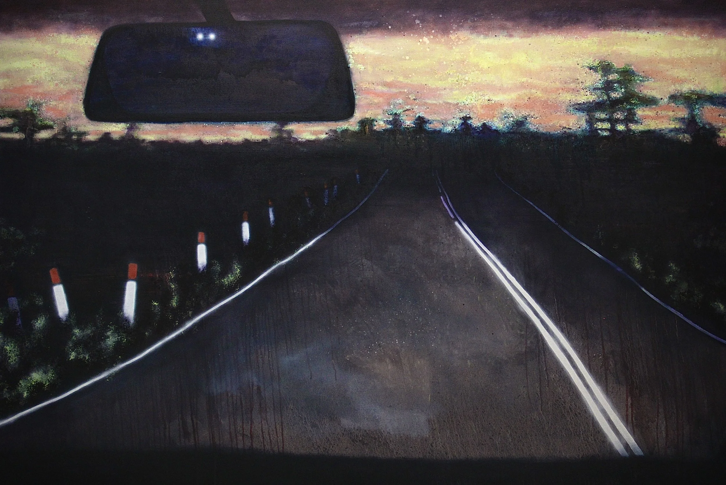 A car's rearview mirror reflecting a colorful sunset sky with trees on the horizon.