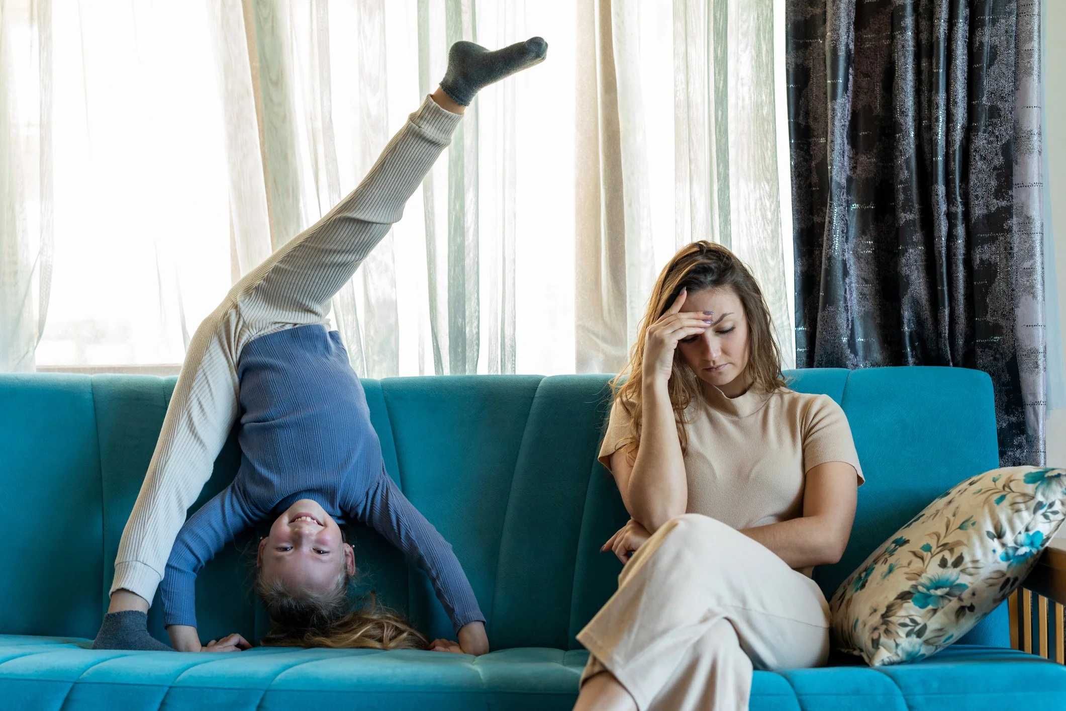 Ein Kind liegt rückwärts auf der Couch, das Kopf hängt nach unten, und ein anderes Kind sitzt auf der Couch, liest ein Buch, während eine Frau neben ihnen sitzt, nachdenklich die Hand an die Stirn legt.