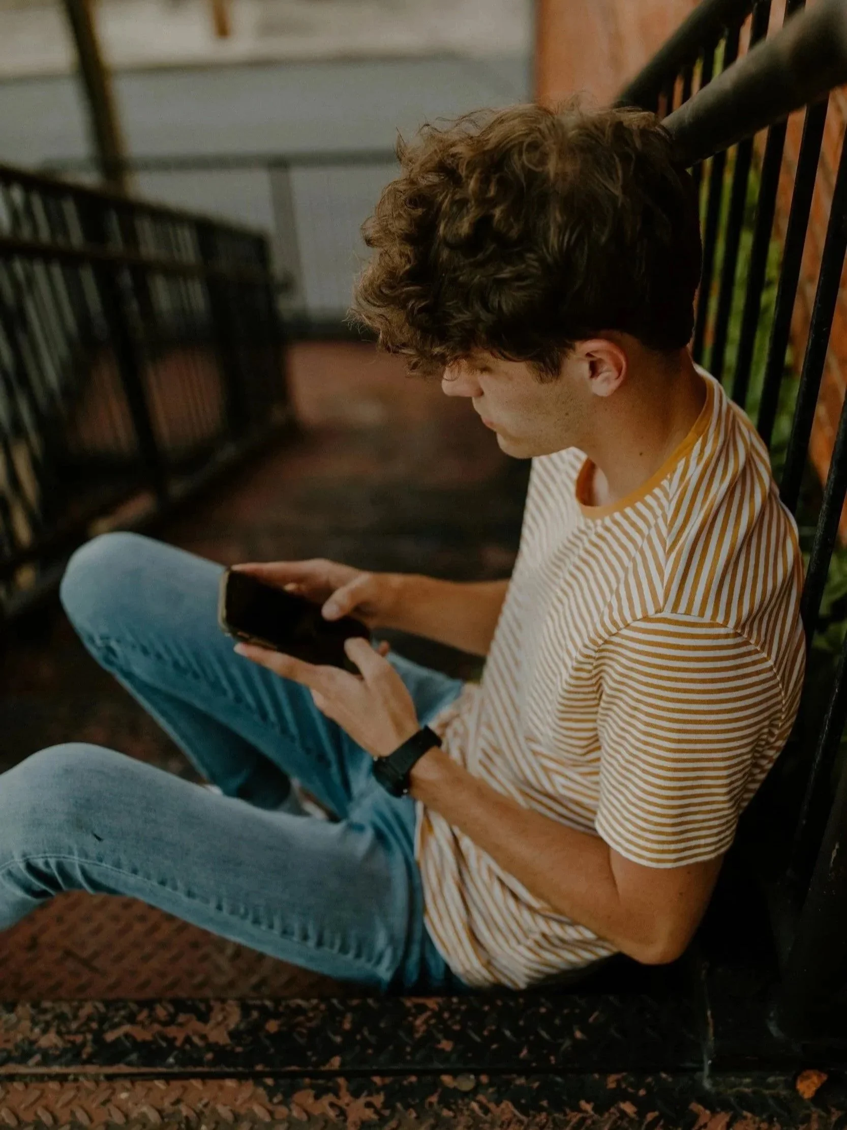 Ein junger Mann sitzt auf einer Bank, trägt ein gelb-weiß gestreiftes T-Shirt und liest auf einem Smartphone.