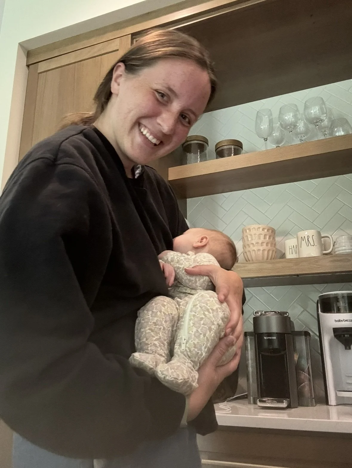A woman holding a sleeping baby in a kitchen. The woman is smiling and looking at the camera. The kitchen has open wood shelves with glasses, mugs, and containers, as well as a coffee maker and other appliances on the counter.