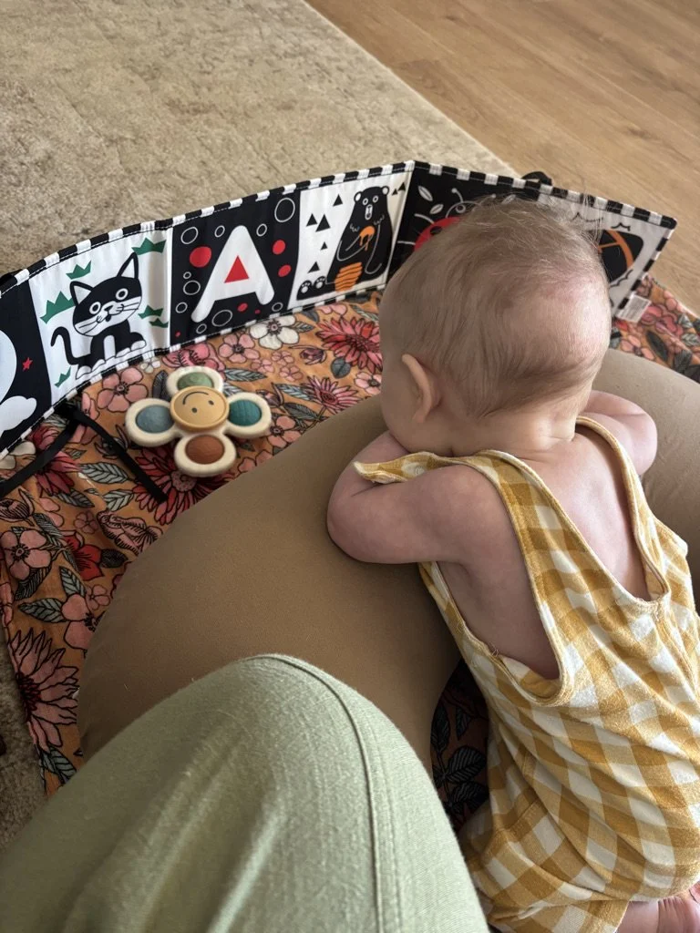 A baby lying on a pillow on a floral blanket, wearing a yellow plaid sleeveless outfit, with a black and white animal-themed playpen in the background and a wooden floor.
