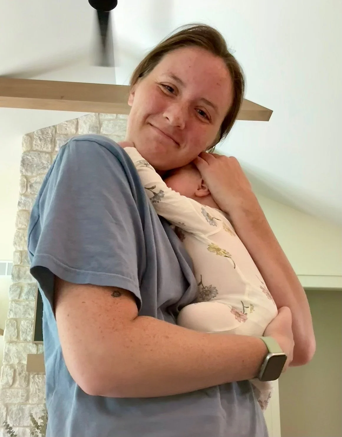 holding a baby close to their chest, with the baby's head resting on their shoulder. The individual is wearing a gray shirt and a smartwatch on their wrist. The background features a light-colored wall, a stone fireplace, and a wooden shelf.