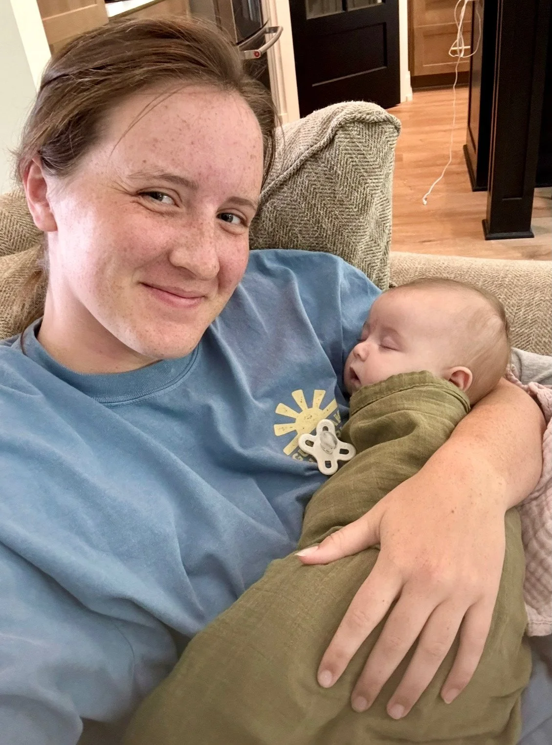 A young woman with brown hair and freckles smiling while holding a sleeping baby on her chest on a beige sofa in a cozy home setting.