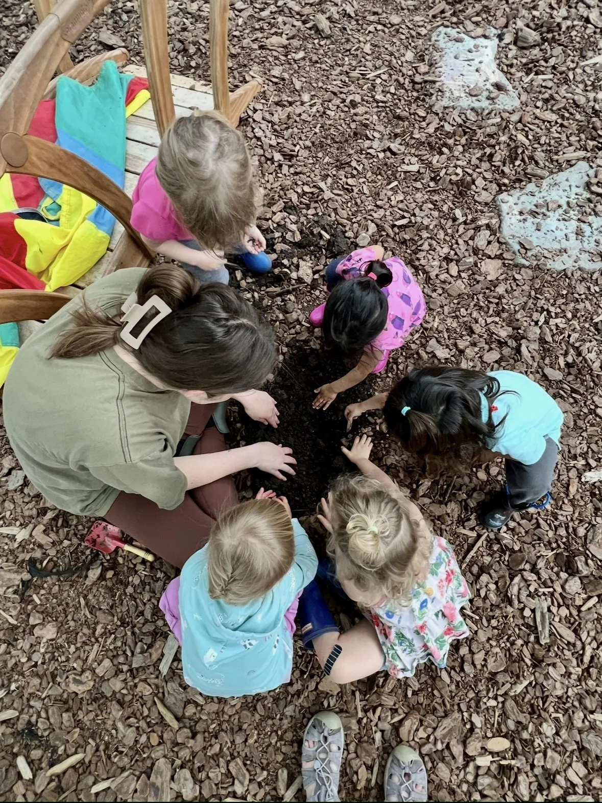 A group of children and an adult sitting on a ground covered in wood chips, digging and exploring in the dirt, with colorful jackets and shoes around.