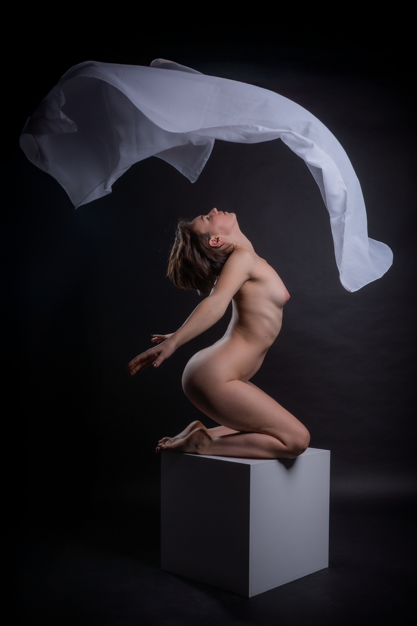 Eine nackte Frau kniet auf einer weißen Plattform, hebt den Oberkörper, während ein weißes Tuch in der Luft über ihr schwenkt, vor einem schwarzen Hintergrund.