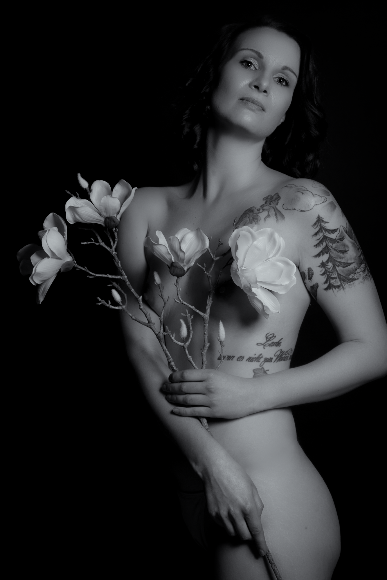 Schwarzweiß-Fotografie einer nackten Frau mit Tattoos, sie hält einen Blumenstrauß mit weißen Blumen vor ihrem Körper, der Hintergrund ist schwarz.
