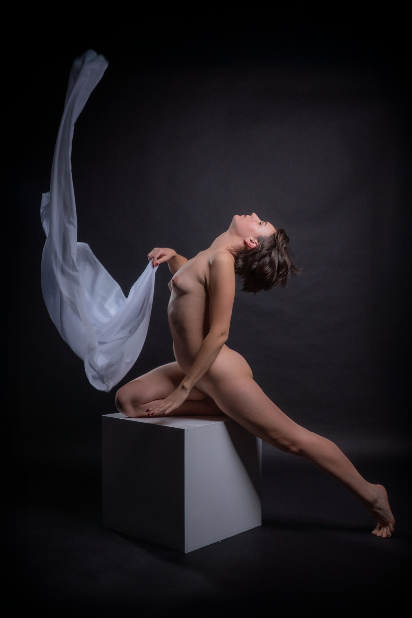 Eine nackte Frau in einer kauernden Pose auf einem weißen Podest, mit schwarzen Hintergrund, hält ein Tuch in der linken Hand, einige Haare sind ungeordnet.