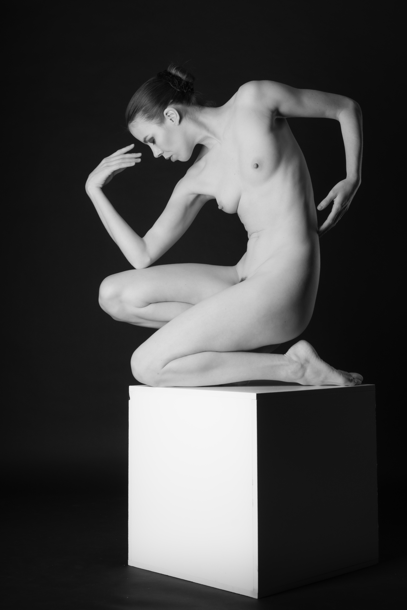 Schwarze und weiße Fotografie einer nackten Frau, die auf einer weißen Plattform sitzt, die in der Mitte des Bildes platziert ist, mit dunklem Hintergrund.