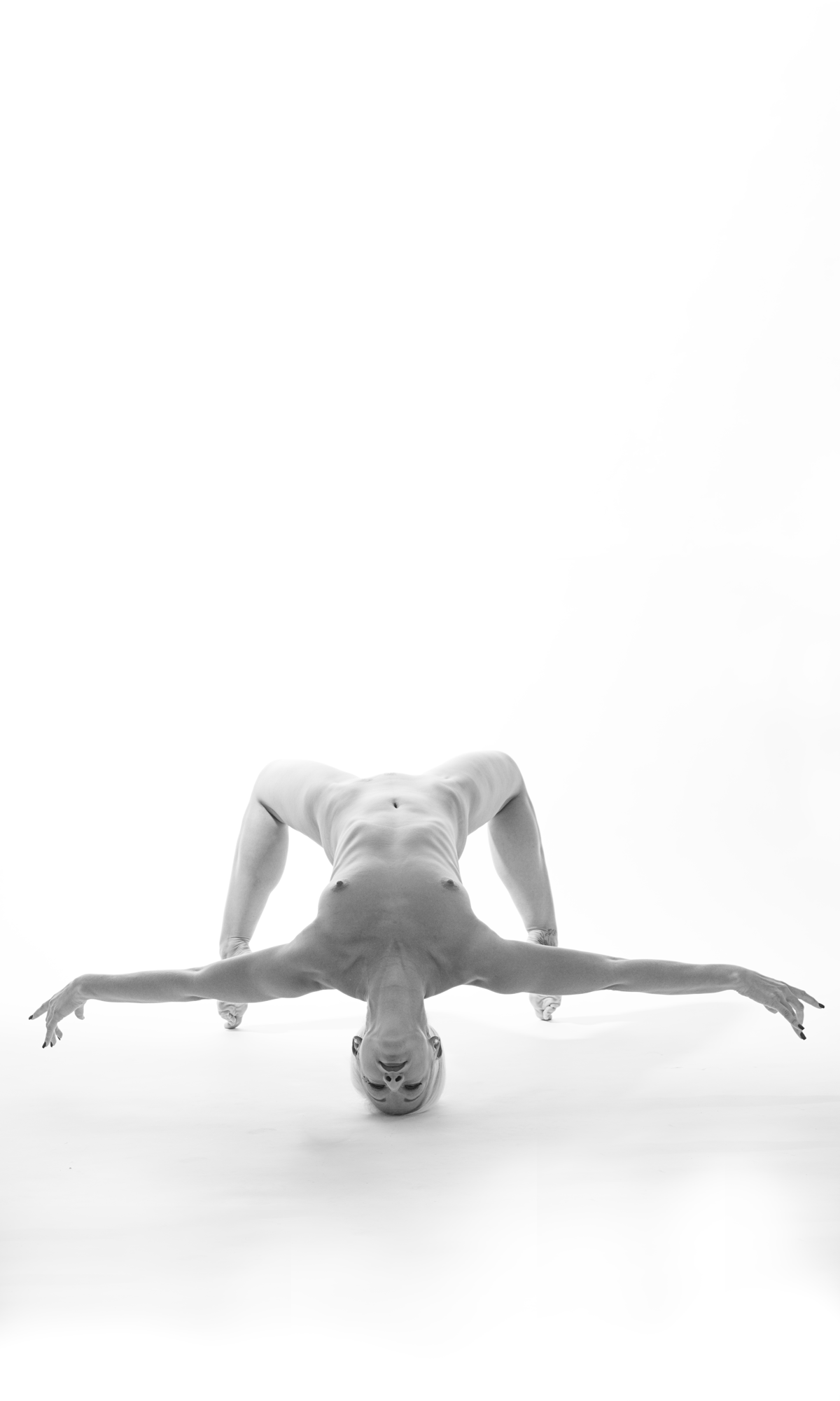 Eine nackte Person macht eine Yoga-Pose auf einer weißen Oberfläche mit einem weißen Hintergrund, die Pose ähnelt der eines umgekehrten V mit Armen und Beinen ausgestreckt.