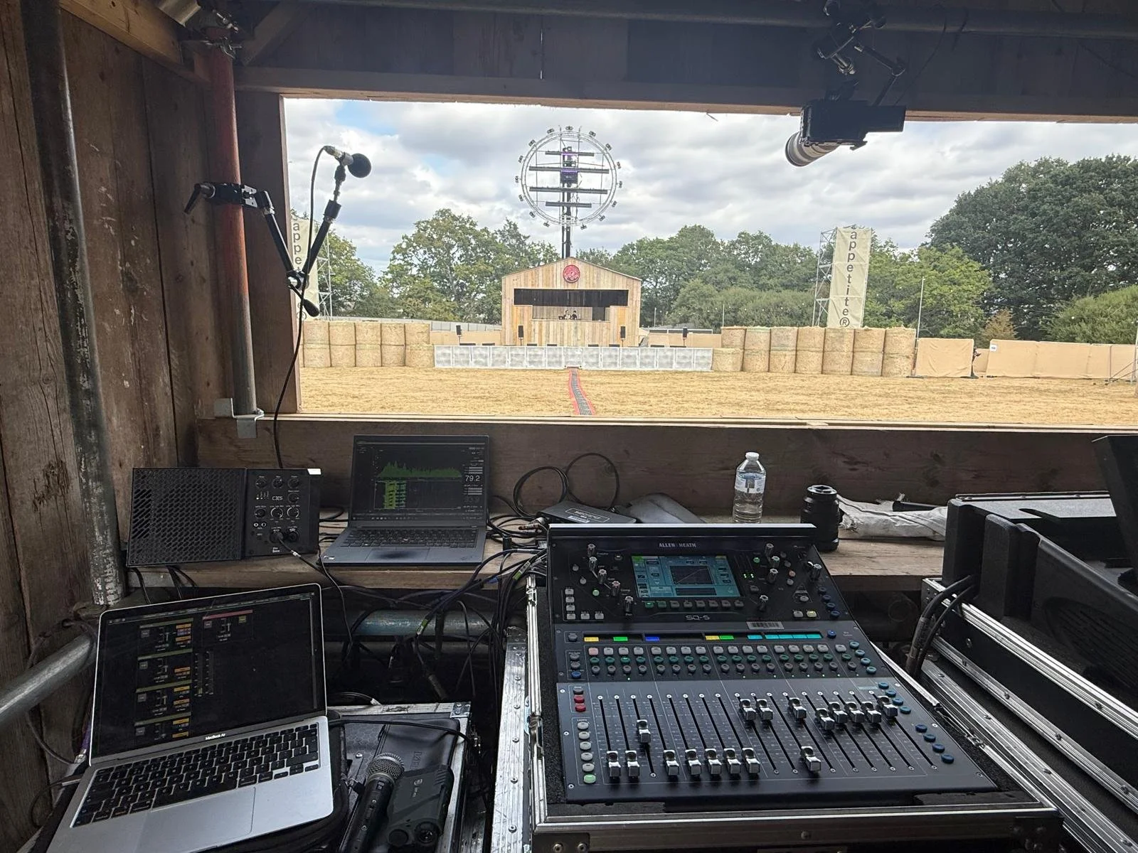 View from a DJ booth overlooking an outdoor concert stage with hay bales and a large circular structural element in the background, set in a rural environment with trees and cloudy sky.
