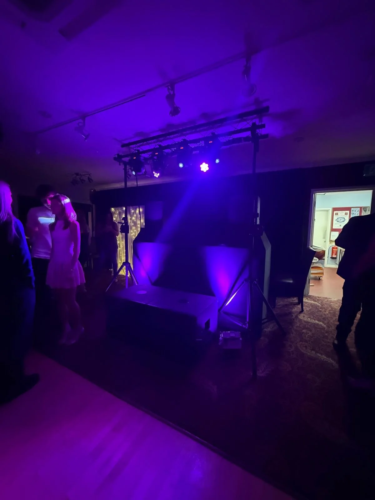 A dimly lit room with purple and blue concert or dance lighting, a DJ booth or stage setup with lighting equipment and speakers, and a few people standing and chatting.