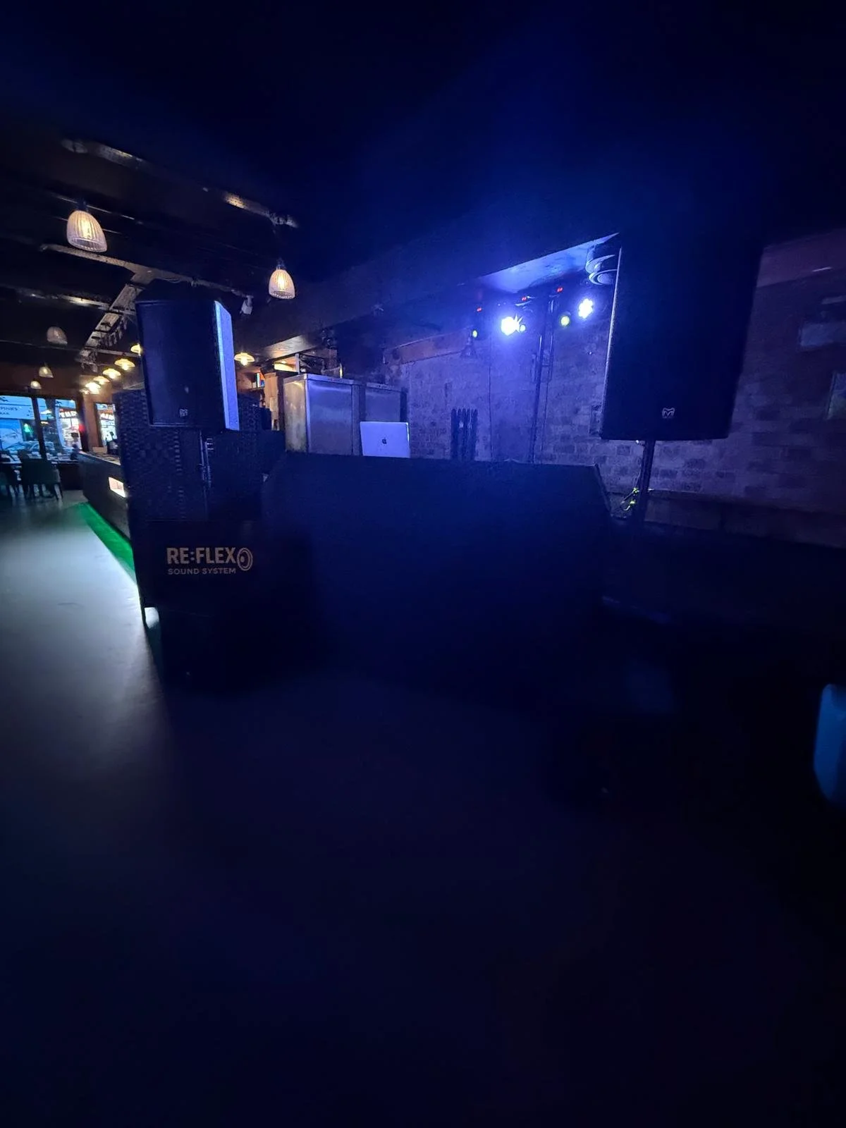 An empty nightclub or bar with DJ equipment including speakers, a laptop, and lighting, with a dark ambiance and some illuminated areas.