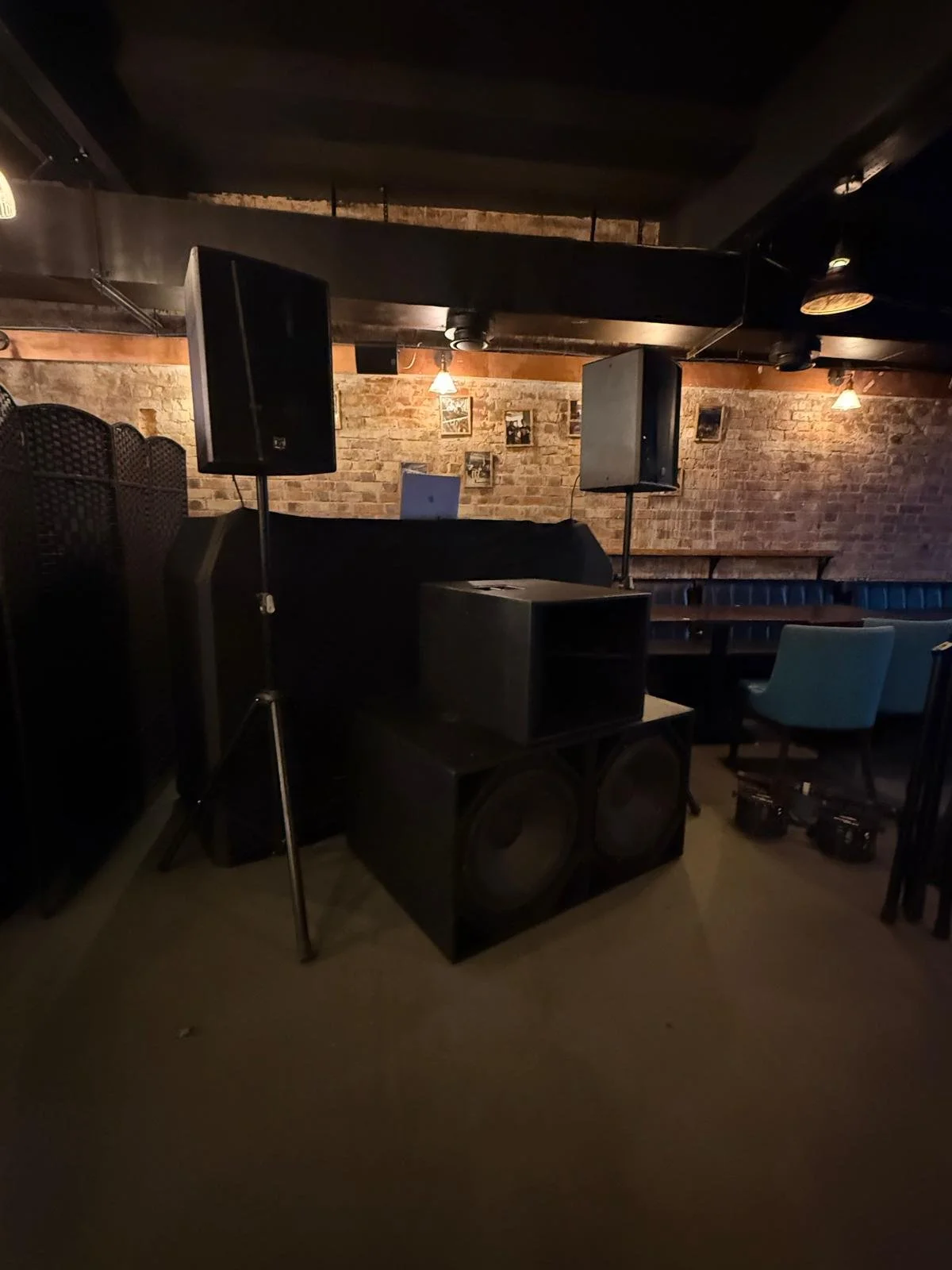 Indoor scene of a dimly lit bar or lounge with speakers, music equipment, brick walls, framed pictures, and seating areas with chairs and booths.