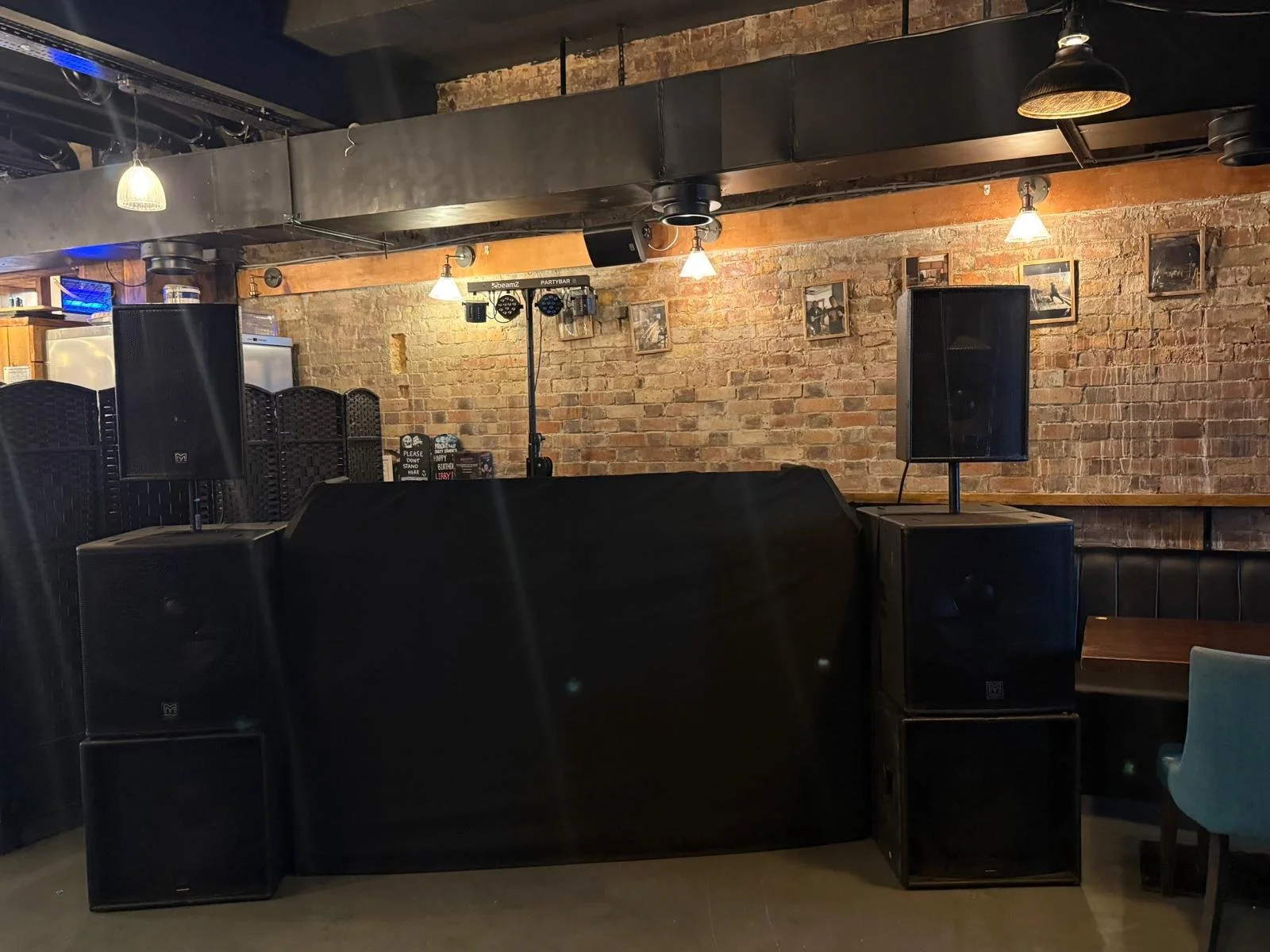 An indoor scene of a bar or restaurant with a brick wall, several framed pictures, and audio equipment including speakers and a DJ setup.