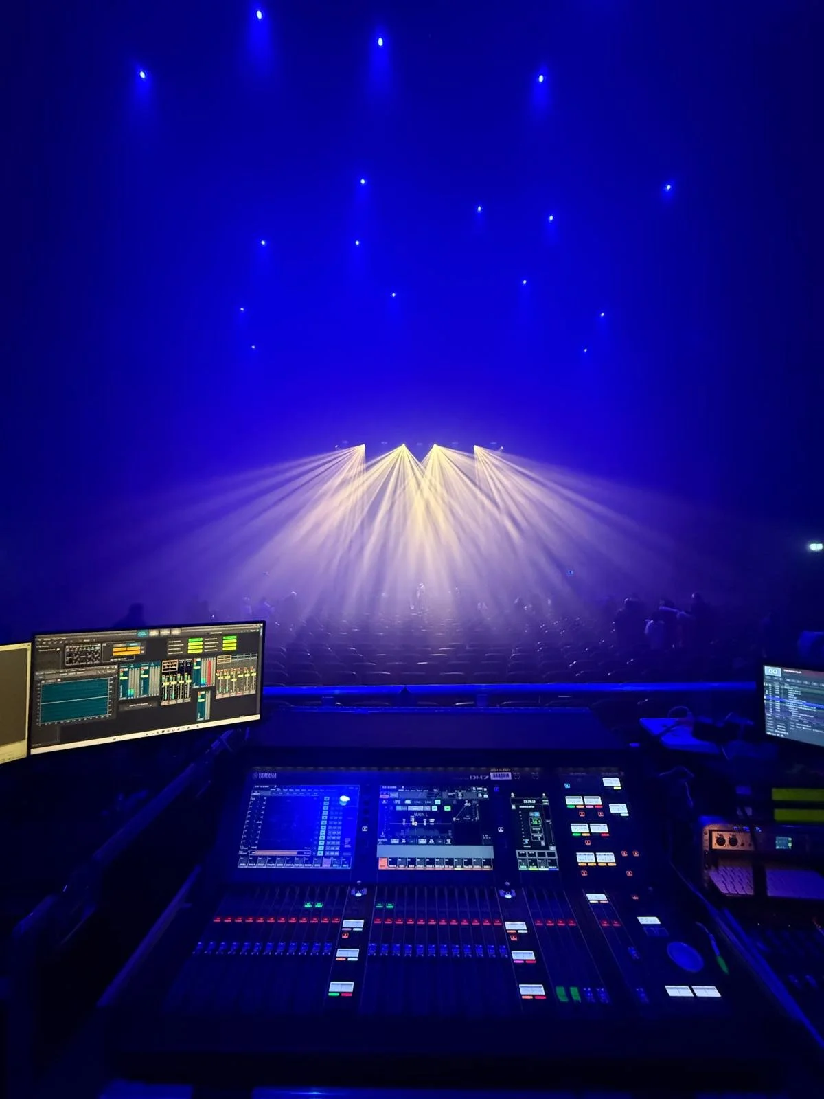 DJ mixing console at a concert with blue and yellow stage lights and empty seats in the background.
