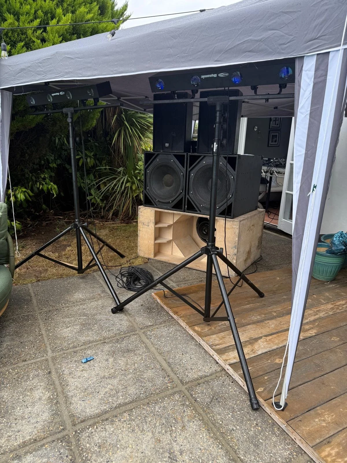 An outdoor DJ setup with large speakers, lighting, and equipment under a canopy, on a patio surrounded by plants.