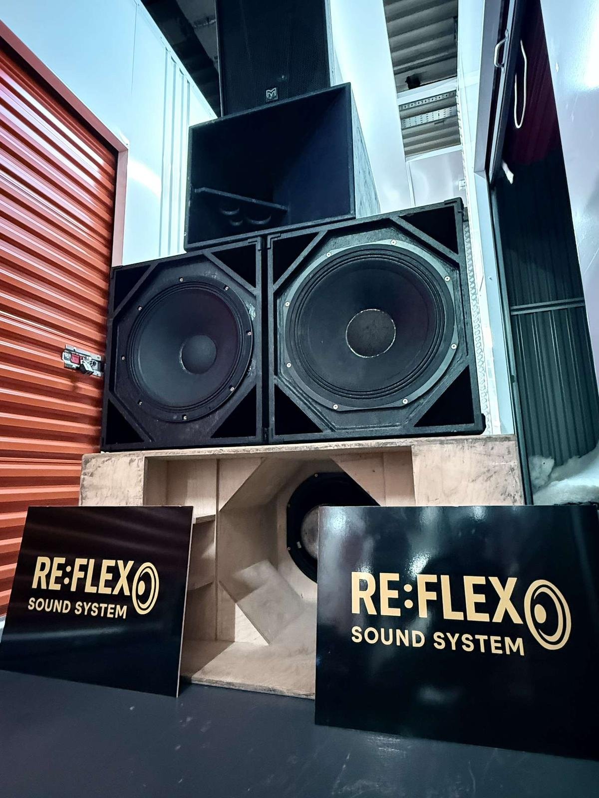 Large professional audio speakers and subwoofers with RE:FLEX SOUND SYSTEM branding on black signs, set up in an indoor storage or display area.
