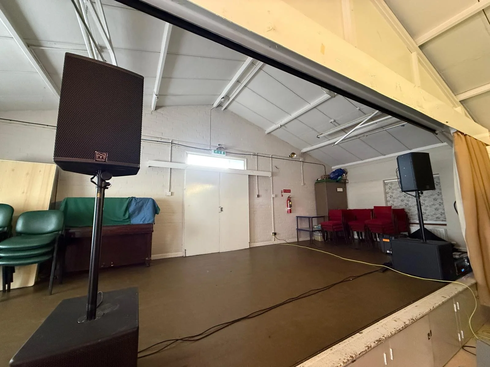 Empty stage with two large speakers, a small table with chairs, and green chairs stacked on a table in a room with white brick walls and a high ceiling.