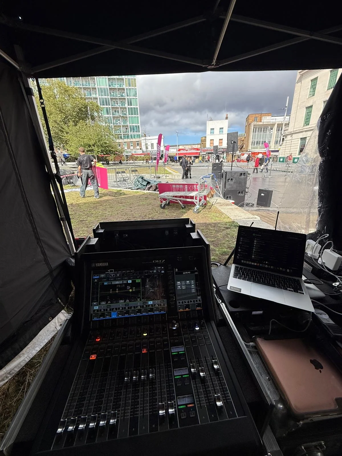 View from inside a tent showing sound mixing equipment and a laptop, looking out onto an outdoor stage setup in a city plaza with people, flags, and tall buildings under cloudy sky.