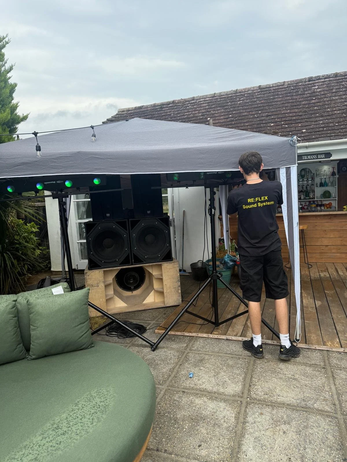 Person setting up a sound system under a canopy outdoors in a backyard, with a sofa in the foreground and a bar area in the background.