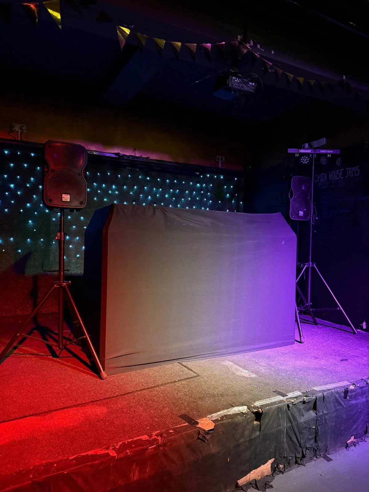 Empty stage with two speakers on stands, a table with a cloth, colorful lights, and decorations hanging from the ceiling, suggesting a performance or event space.