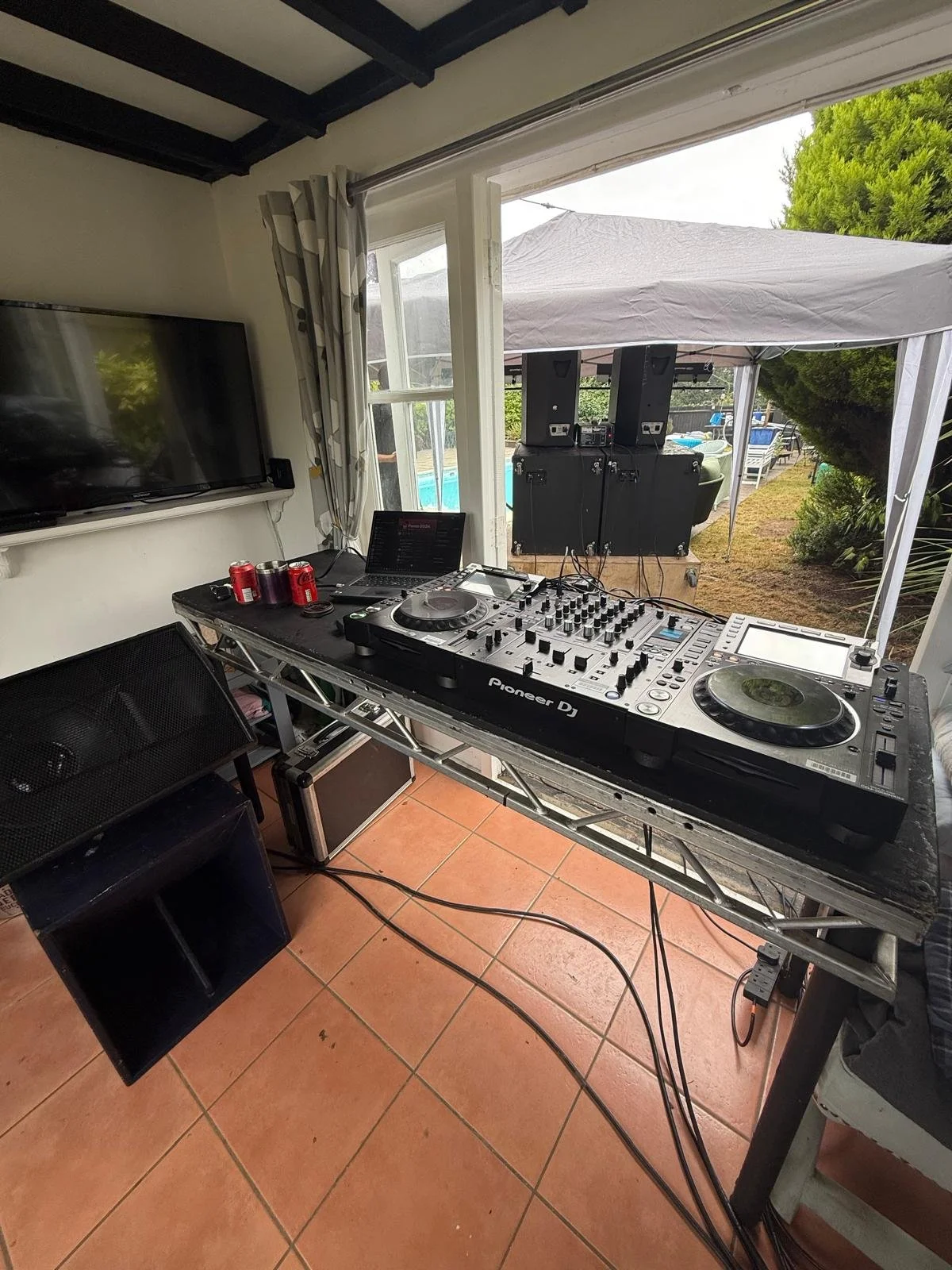 A DJ setup with a Pioneer DJ controller, laptop, soda cans, and speakers outside under a canopy, with a pool and greenery in the background.