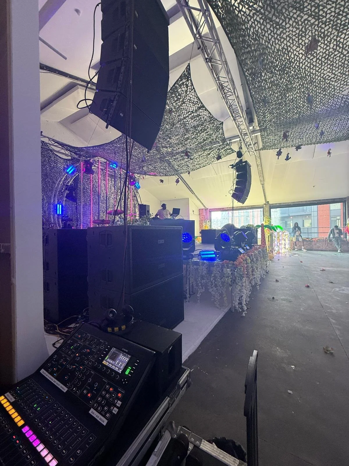 Music event stage with DJ setup, large speakers, lighting equipment, and decorative floral arrangements, under a camouflage net ceiling with people in the background.