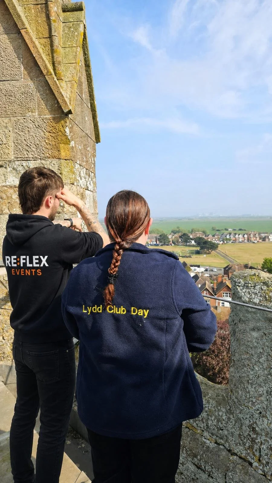 Two people standing on a viewing platform overlooking a scenic landscape of houses, trees, and fields, with a stone wall on their left and a partly cloudy sky above.