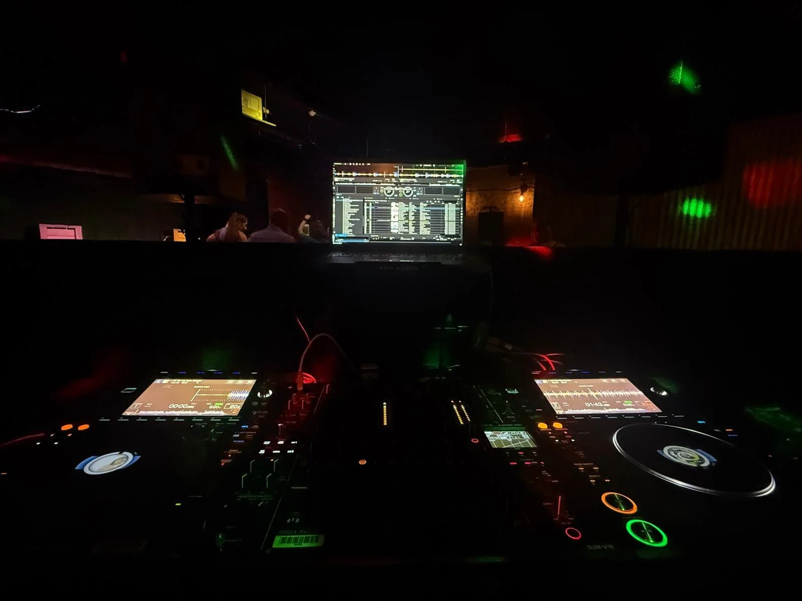 A dark club scene featuring DJ equipment with colorful LED lights, a laptop displaying music software, and a DJ mixing tracks while people dance in the background.