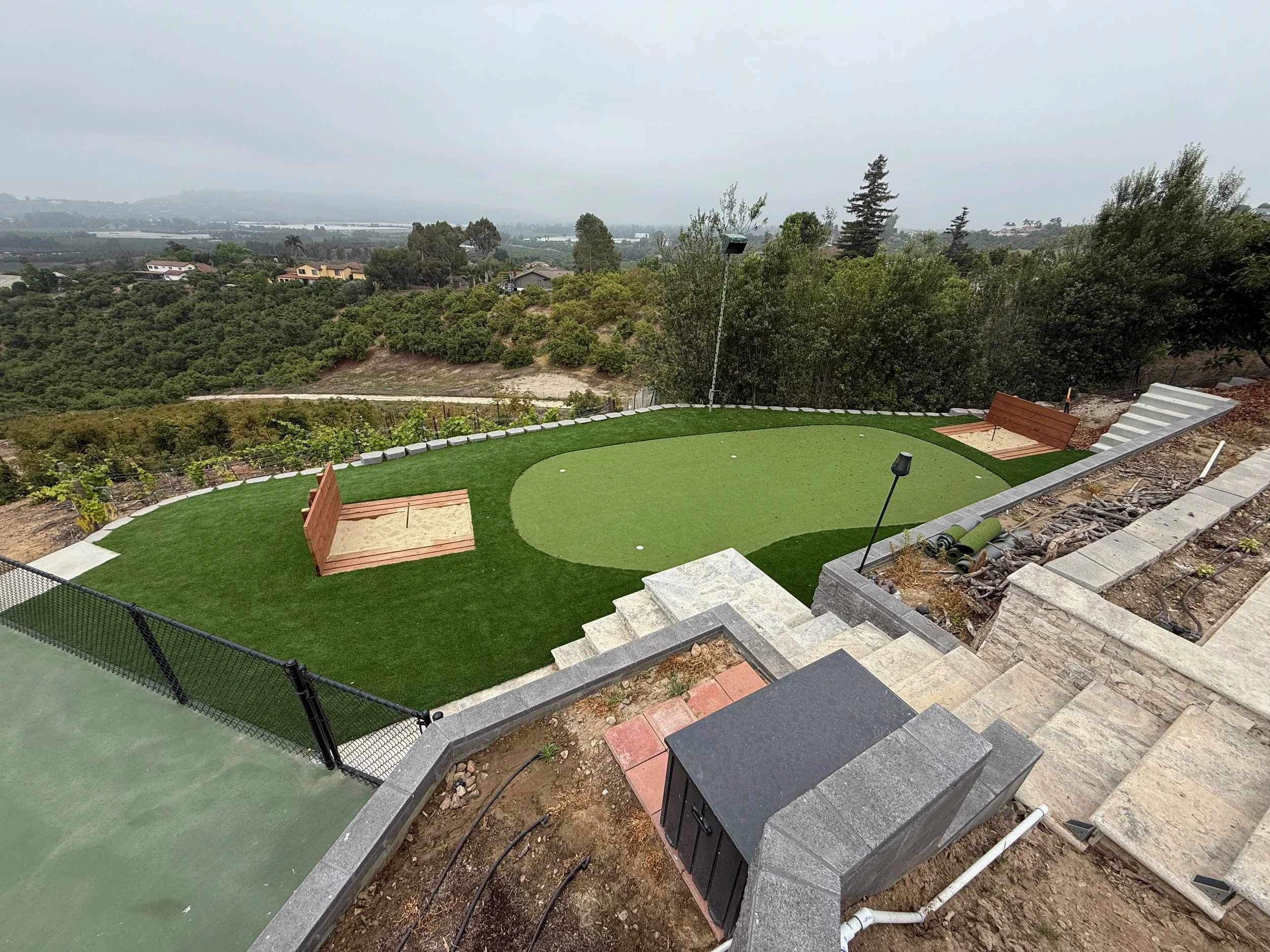 A backyard with artificial grass, a putting green, benches, and a retaining wall with stairs, overlooking a hilly landscape with trees and houses under an overcast sky.