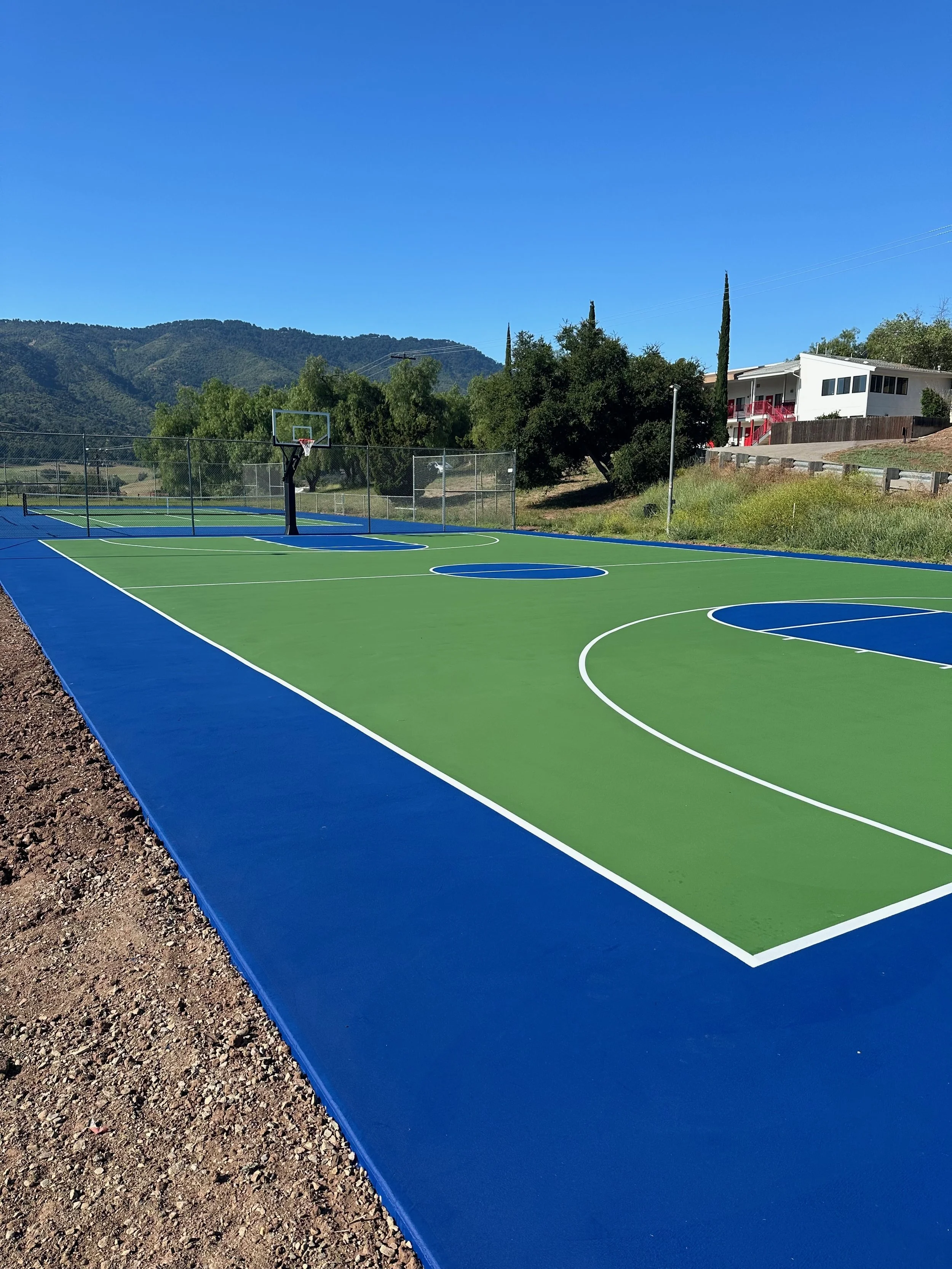 A newly painted outdoor basketball court with bright blue and green surface, surrounded by a chain-link fence and scenic mountain backdrop under clear blue sky.