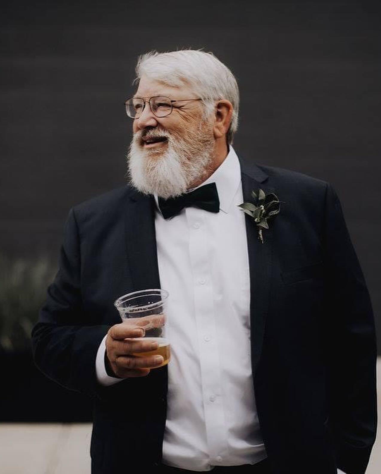 An elderly man with white hair and beard wearing glasses, a black tuxedo, and a bowtie, holding a glass of beer and smiling.
