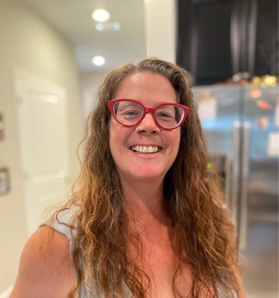 A woman with curly brown hair, wearing red glasses and a light-colored top, smiling in a home kitchen.