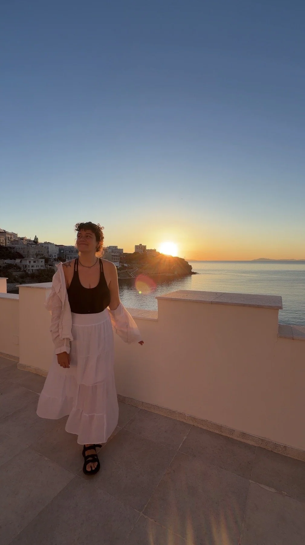 A young woman walking on a terrace at sunset, overlooking the ocean with houses on a hillside in the background. She wears a black top, a long white skirt, and sandals, smiling softly as the sun sets behind her.