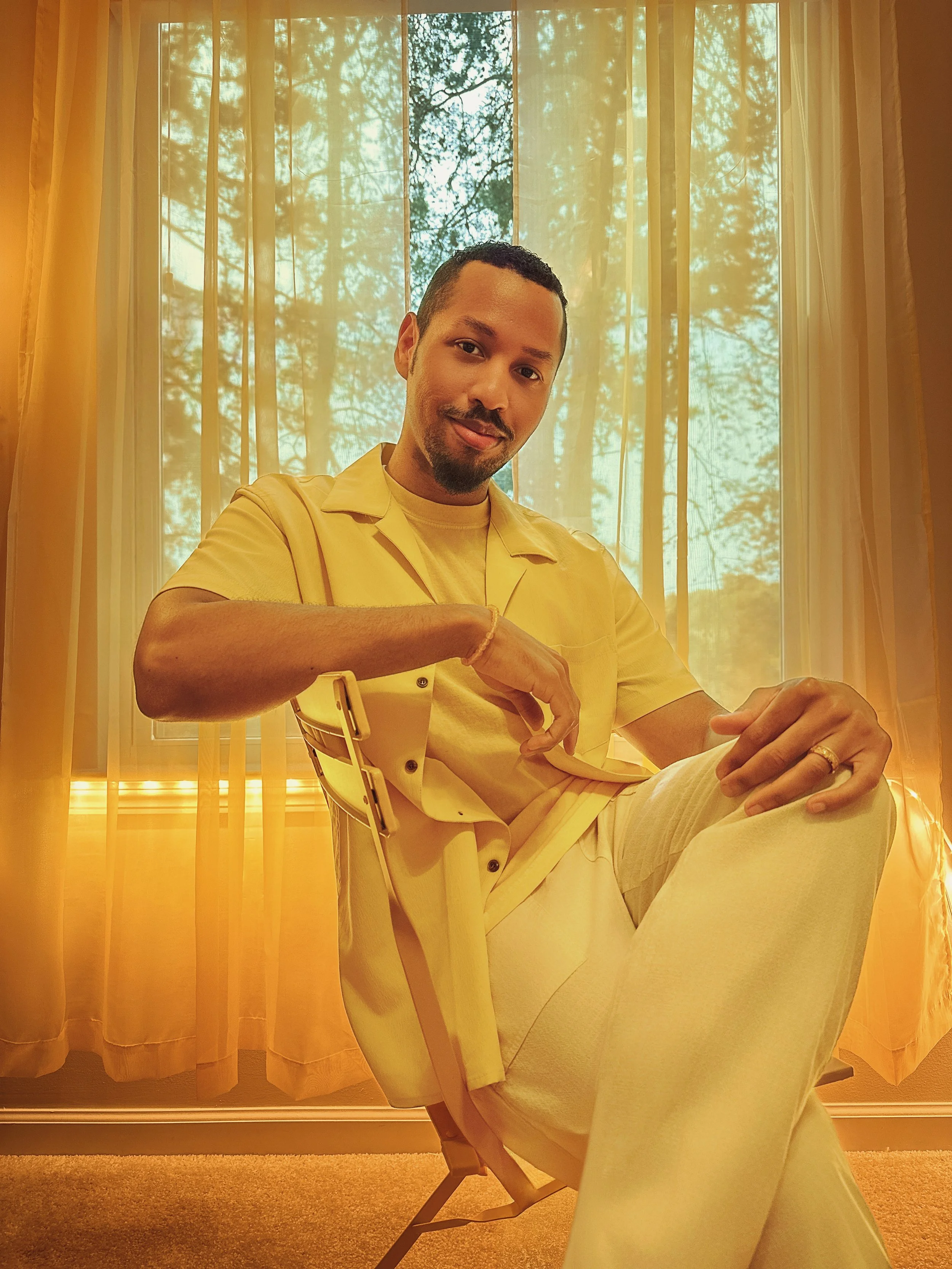 The Doctor in Yellow's founder and owner, Keiraj Gillis, sitting in a room in a yellow chair wearing a yellow collared shirt and white pants in front of sheer yellow curtains covering a window with a view of trees.