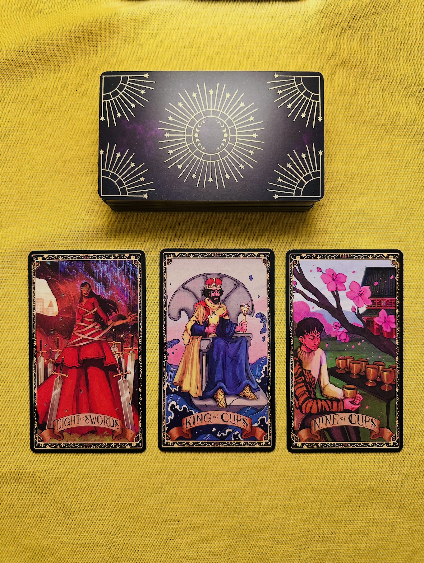 Three cards from The Cosmic Wisdom Tarot deck - the Eight of Swords, the King of Cups, and the Nine of Cups - laid out in a row on a yellow cloth with a deck above them that has a black and purple background with a gold-foiled sun pattern.