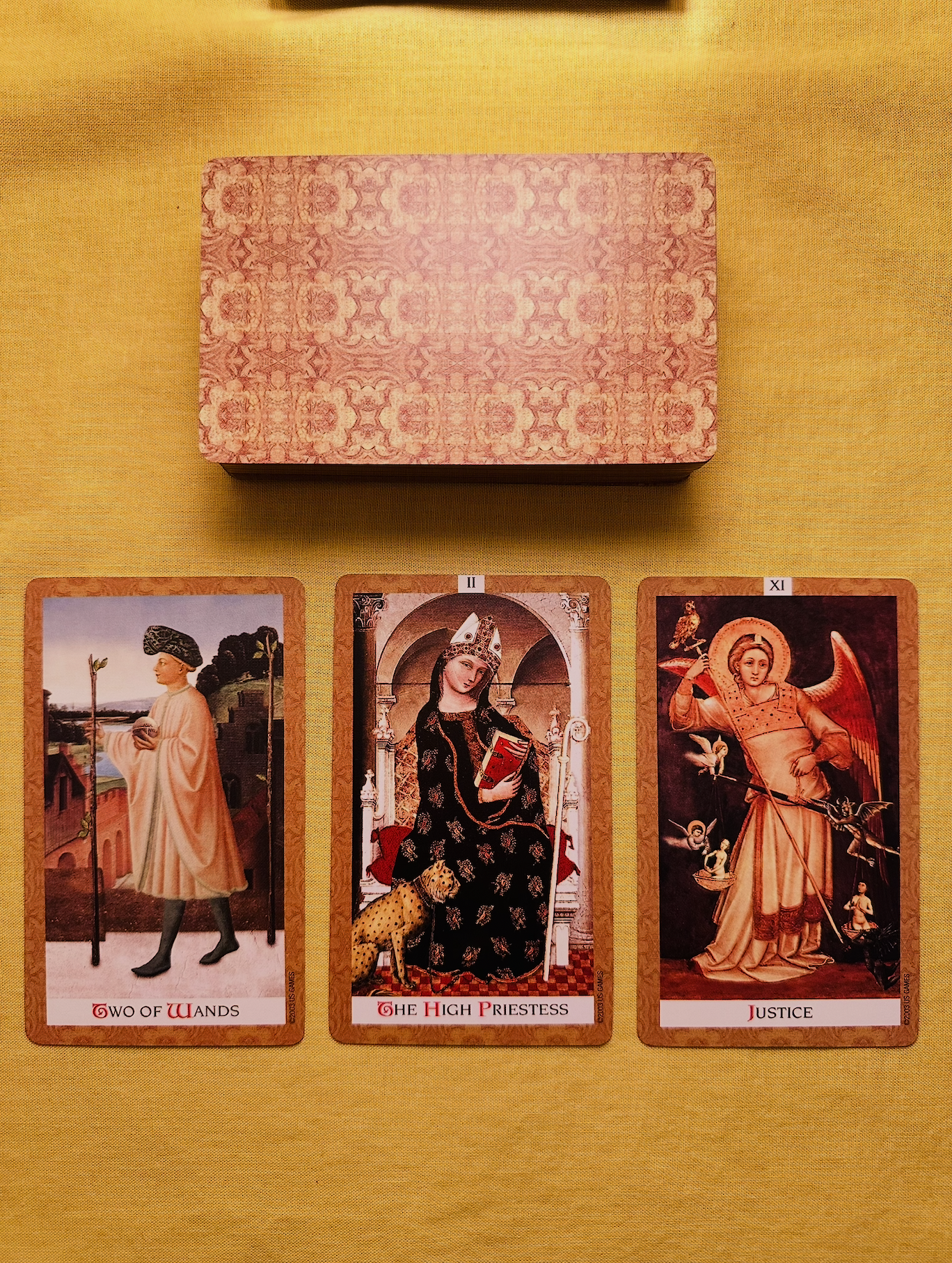 Three cards from the Golden Tarot deck - the Two of Wands, The High Priestess, and Justice - laid out in a row on a yellow cloth with a deck above them that has an ornate gold and brown pattern.