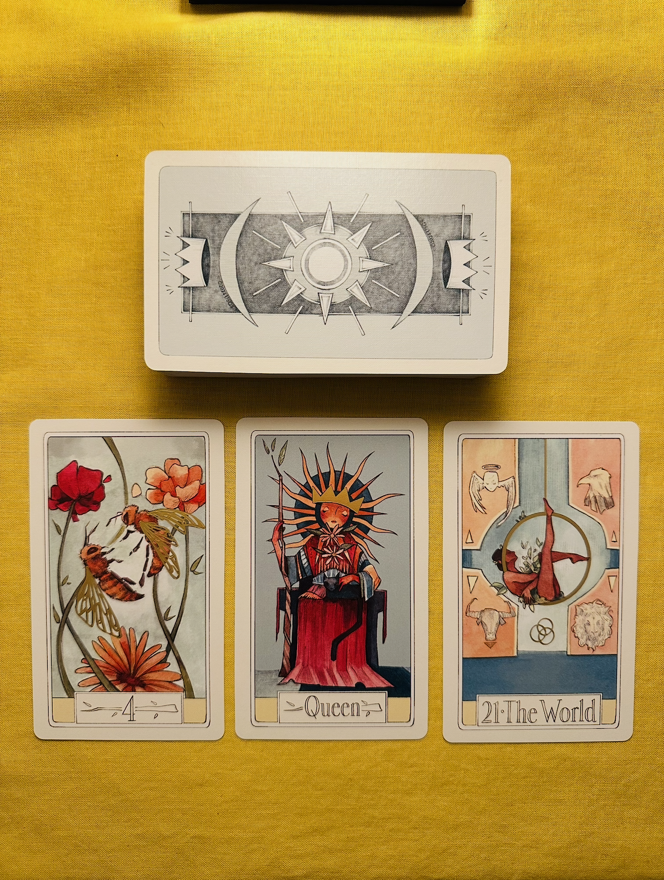 Three cards from the Curious Travels Tarot deck - the Four of Wands, the Queen of Wands, and the The World - laid out in a row on a yellow cloth with a deck above them that has a light blue background with a crown and crescent moon pattern.