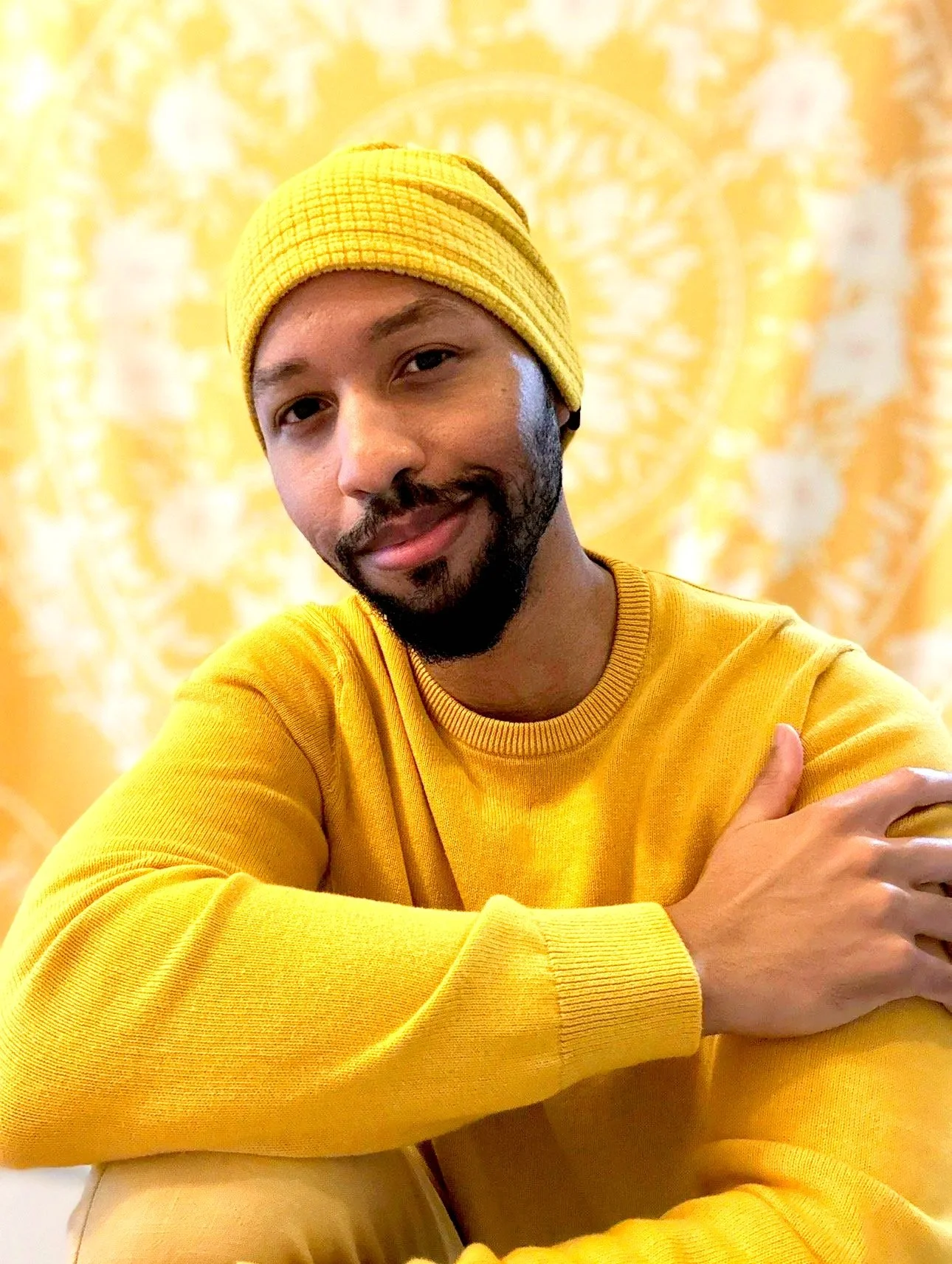 The Doctor in Yellow's founder and owner, Keiraj Gillis, wearing a bright yellow long-sleeved shirt and a bright yellow beanie in front of a yellow and white patterned background.