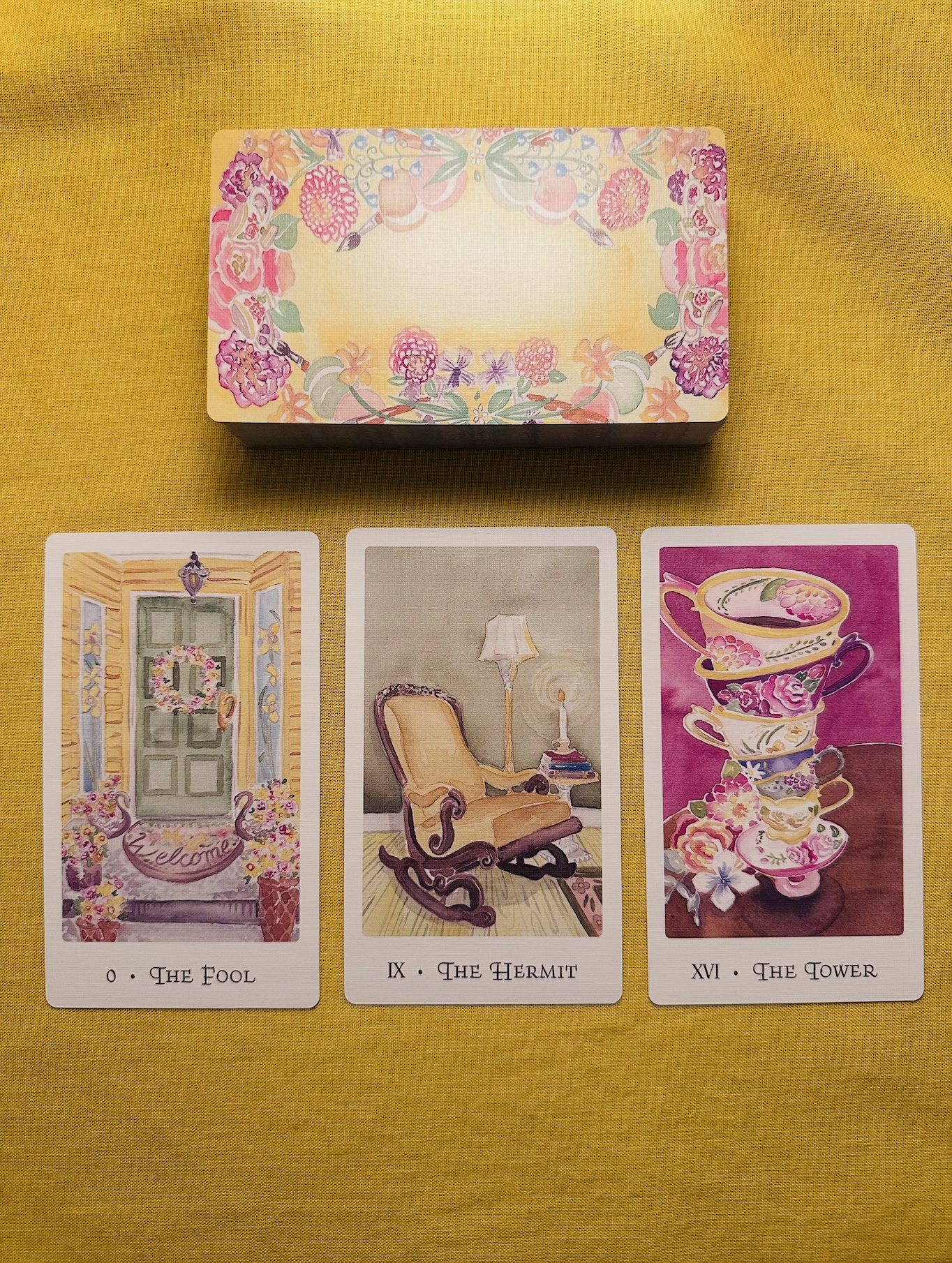 Three cards from the Yellow House Tarot deck - The Fool, The Hermit, and The Tower - laid out in a row on a yellow cloth with a deck above them that has a background with pastel yellow and pink patterns.