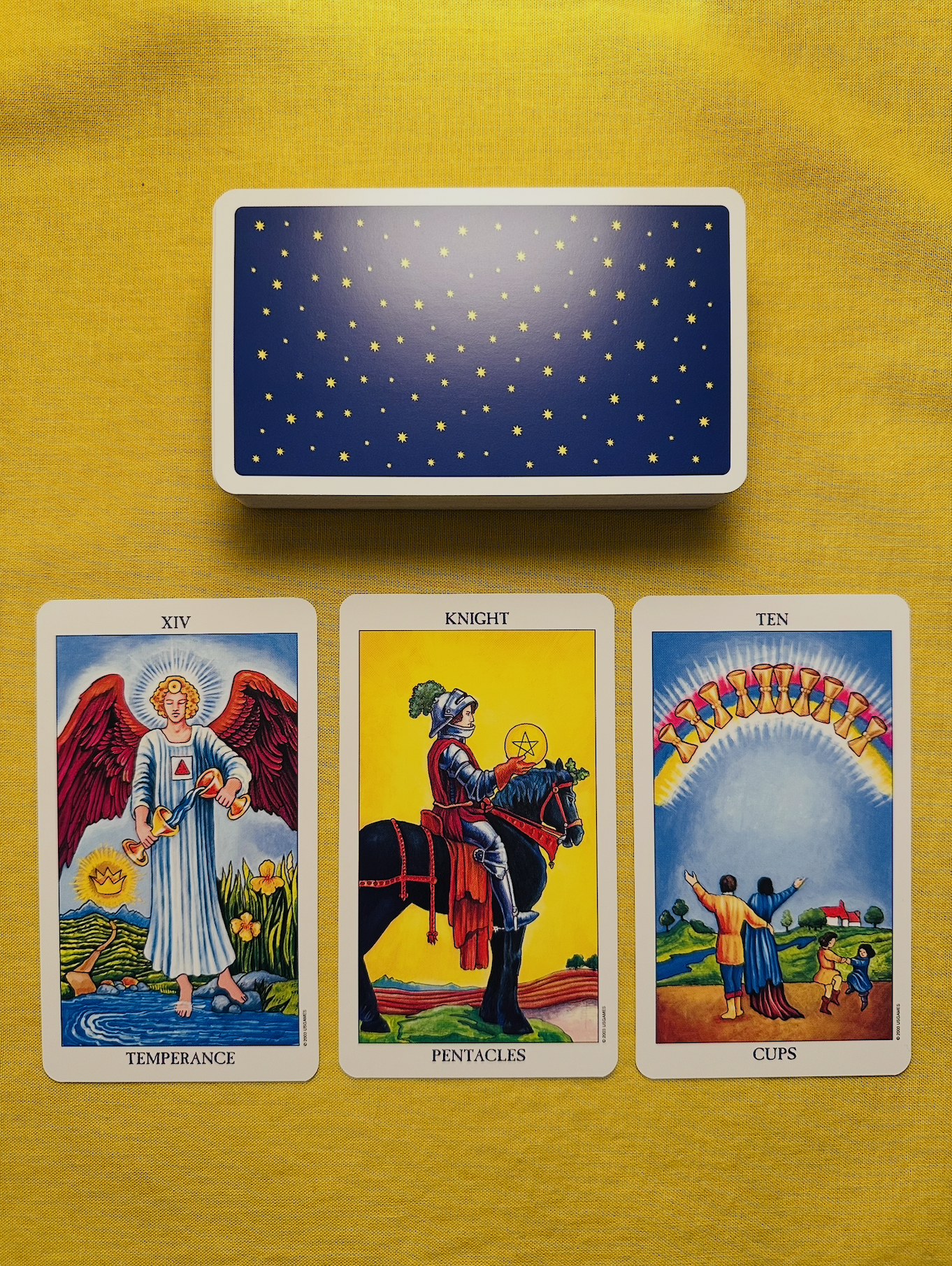 Three cards from the Radiant Waite-Smith deck - Temperance, the Knight of Pentacles, and the Ten of Cups - laid out in a row on a yellow cloth with a deck above them that has a dark blue starry patterned background.